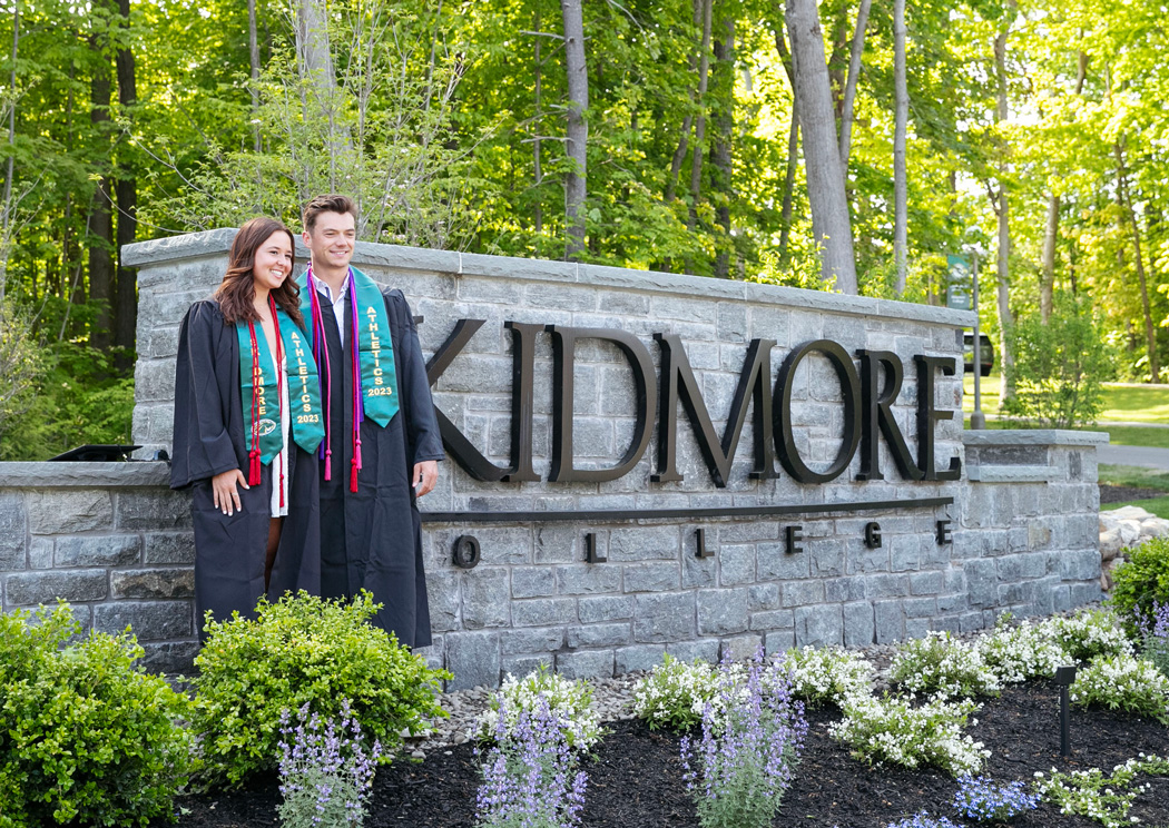 two college graduates post in front of a campus entrance sign that says ¼ϲʿ